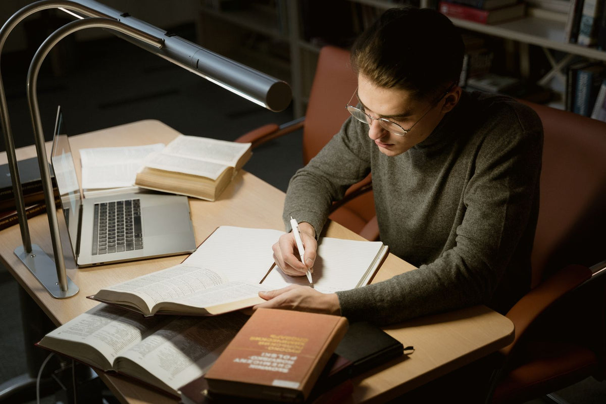 Young adult studying with books and a laptop, taking notes at a desk under a lamp - how to study after work when you are tired